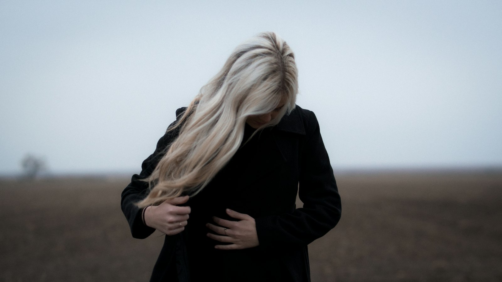 A woman in a black coat standing in an open field under a gray sky